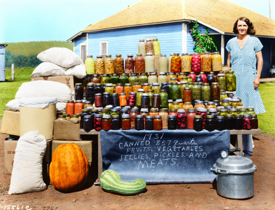A woman in a blue gingham dress stands next to a display of canned fruits, vegetables, and meats outside. Jars are stacked on a table with a sign reading, "1931 Canned 857 quarts fruits, vegetables, jellies, pickles and meats.