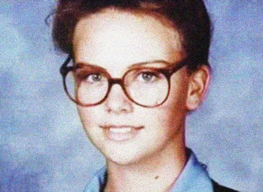 A school portrait of a young person with short brown hair, wearing large round glasses and a blue collared shirt, smiling slightly in front of a mottled blue background.