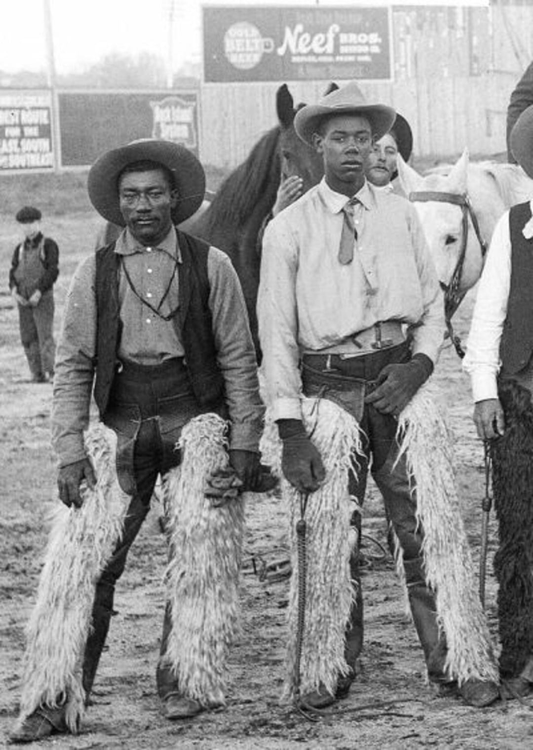 Two Black cowboys wearing hats, shirts, vests, neckerchiefs, and chaps stand side by side outdoors with horses in the background at what appears to be a rodeo or fairground.