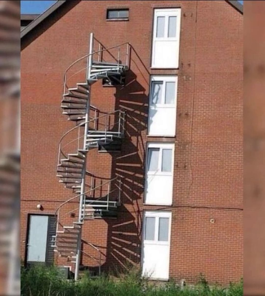A metal spiral staircase is attached to a brick building, leading to doors on upper floors. The doors open to nowhere, with no balconies or landings, making the staircase appear unusable and unsafe.