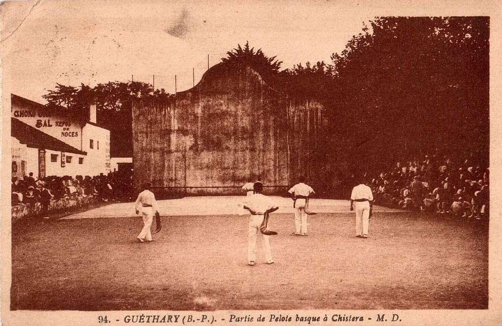 Vintage postcard showing four men in white uniforms playing Basque pelota with chistera baskets on an outdoor court, watched by spectators seated along the sides in front of a tall walled backdrop.