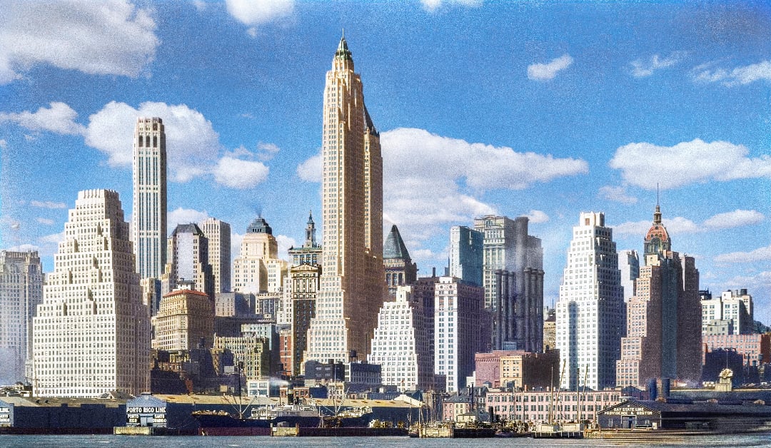 Colorful vintage photo of a city skyline with tall skyscrapers, blue sky, and scattered clouds. The buildings have varied architectural styles, and there are docks with warehouses in the foreground.