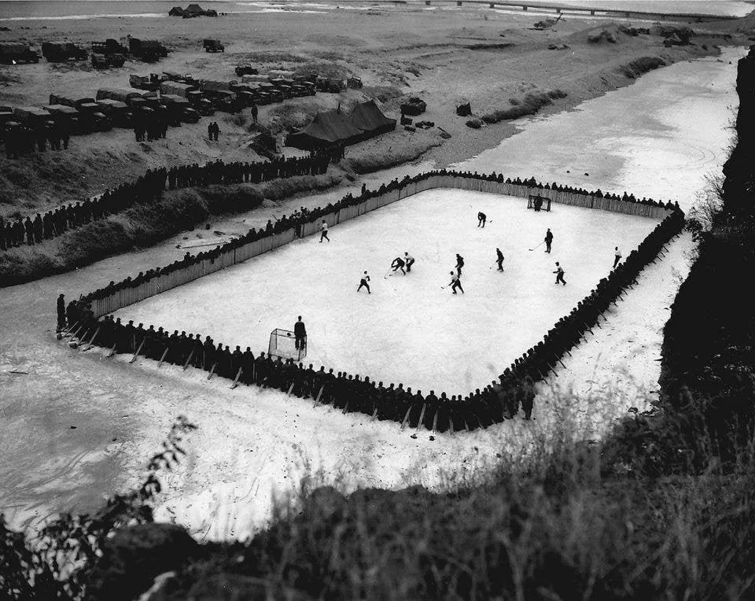 An aerial view of a group of people playing ice hockey on an outdoor rink surrounded by spectators, with vehicles and tents in the background along a frozen river.