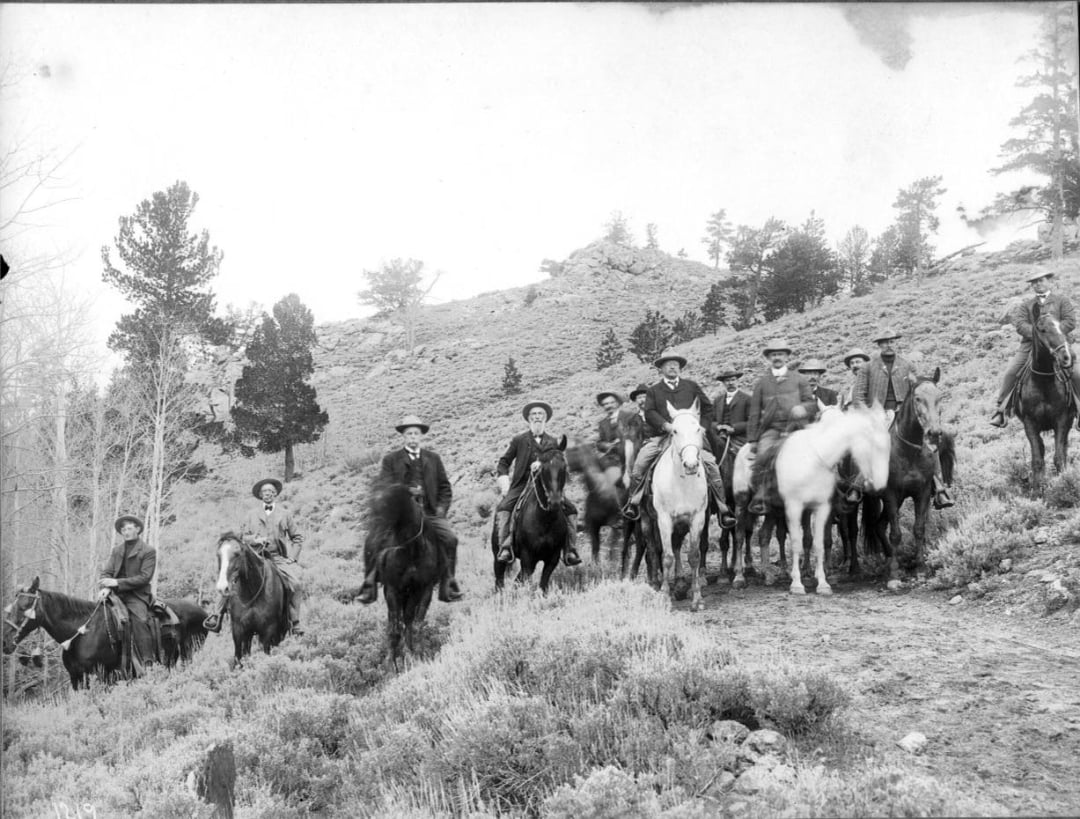 A group of people ride horses through a hilly, grassy landscape with trees and a rocky slope in the background. The riders, wearing hats and outdoor clothing, are positioned in a loose formation facing the camera.
