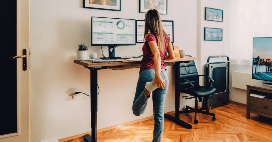 A person stands at a height-adjustable desk, working on dual monitors while stretching one leg behind them in a home office with wooden floors and framed pictures on the wall.