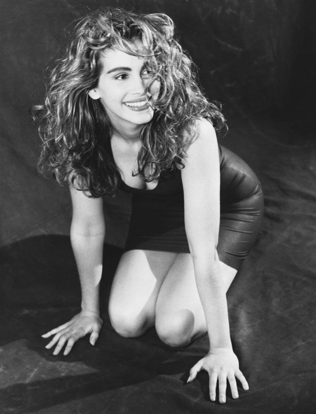 A woman with long, wavy hair kneels on the ground, smiling brightly. She wears a sleeveless, fitted dress and looks off to the side. The image is in black and white.