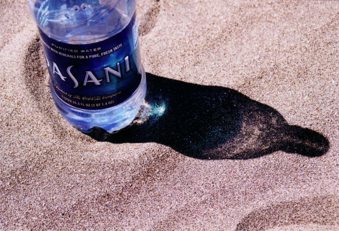 A plastic Dasani water bottle sits upright on sandy ground, casting a dark, crisp shadow to the right on the textured sand.