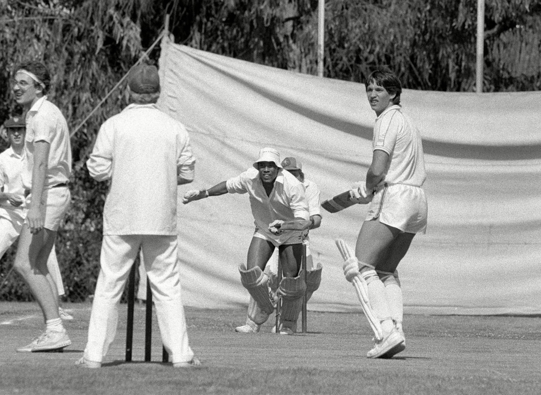Black and white photo of a cricket match in progress; players in white uniforms are on the field, with one wicketkeeper crouching behind the stumps and two batsmen at the crease. Trees and a fabric screen are in the background.