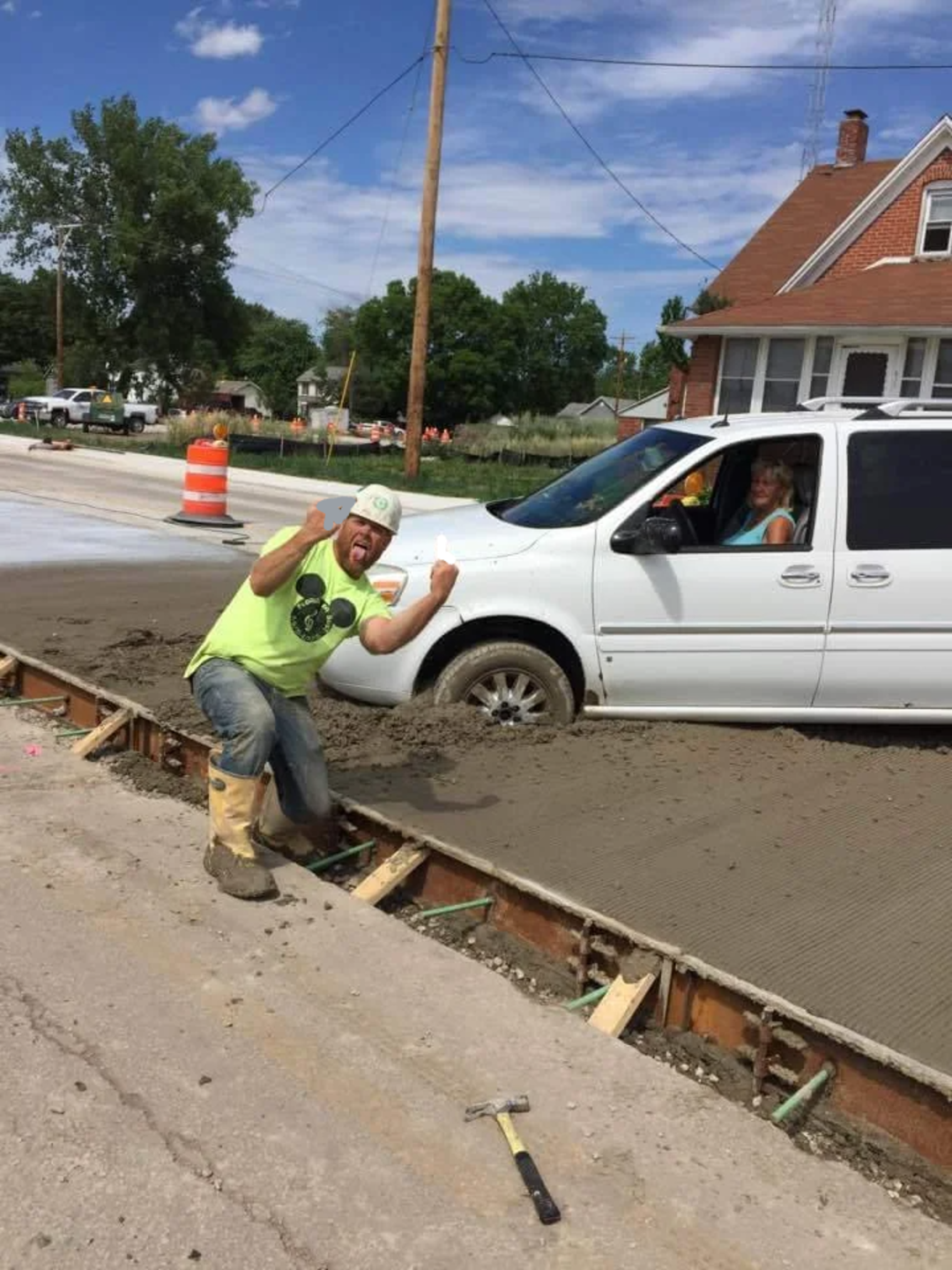 A man in a neon shirt and white hard hat gestures in frustration at a white SUV stuck in freshly poured concrete on a road construction site, while a woman sits in the driver's seat.