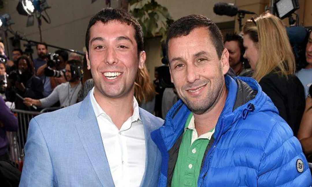 Two men smile for a photo at an outdoor event. One wears a light blue suit, and the other wears a blue puffer jacket over a green polo. People and cameras are visible in the background.