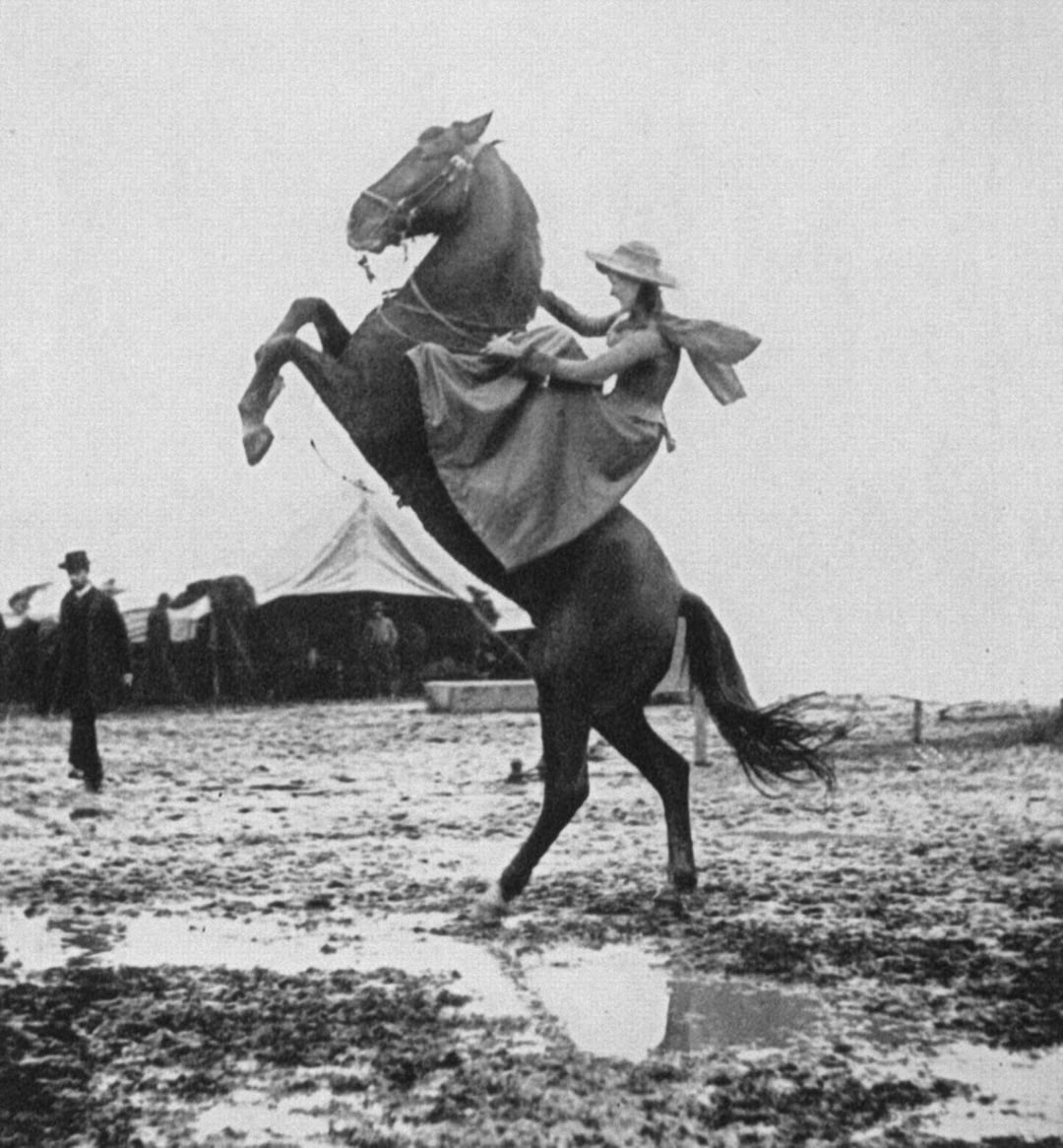 A woman in a dress and wide-brimmed hat rides a rearing horse near a circus tent on muddy ground, while a man in a top hat walks nearby.