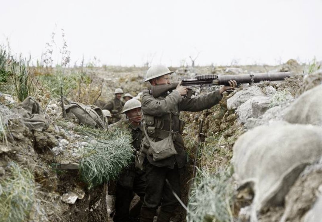 A group of World War I soldiers in helmets stand in a muddy trench; one soldier aims a machine gun while others watch or crouch behind him. Grassy earth and sandbags are visible around the trench.