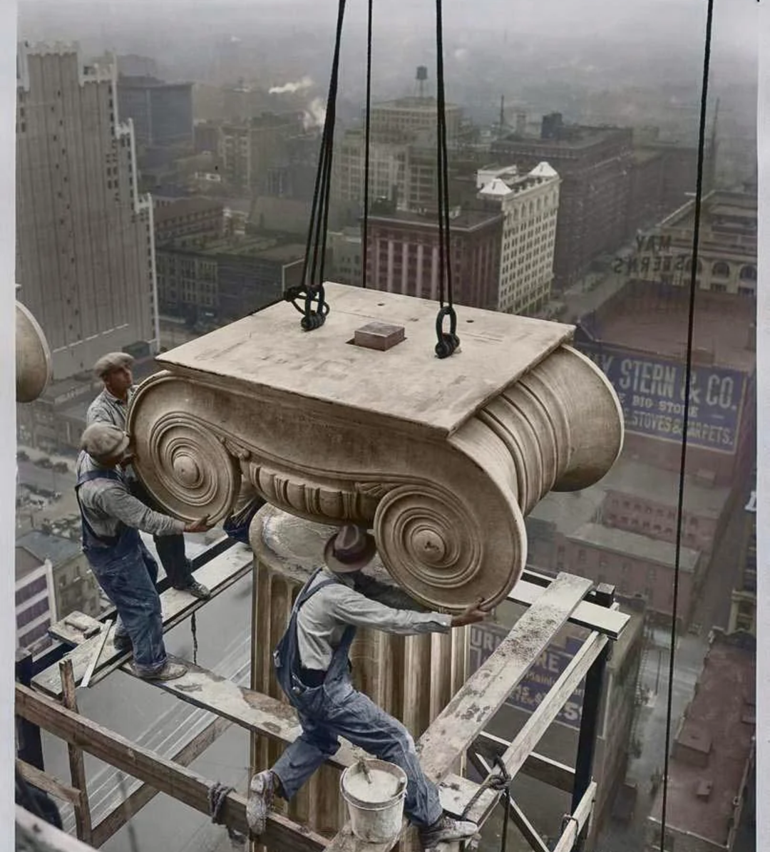 Three construction workers guide a large classical column capital being hoisted by crane atop a high-rise building, with a foggy cityscape and tall buildings visible in the background.