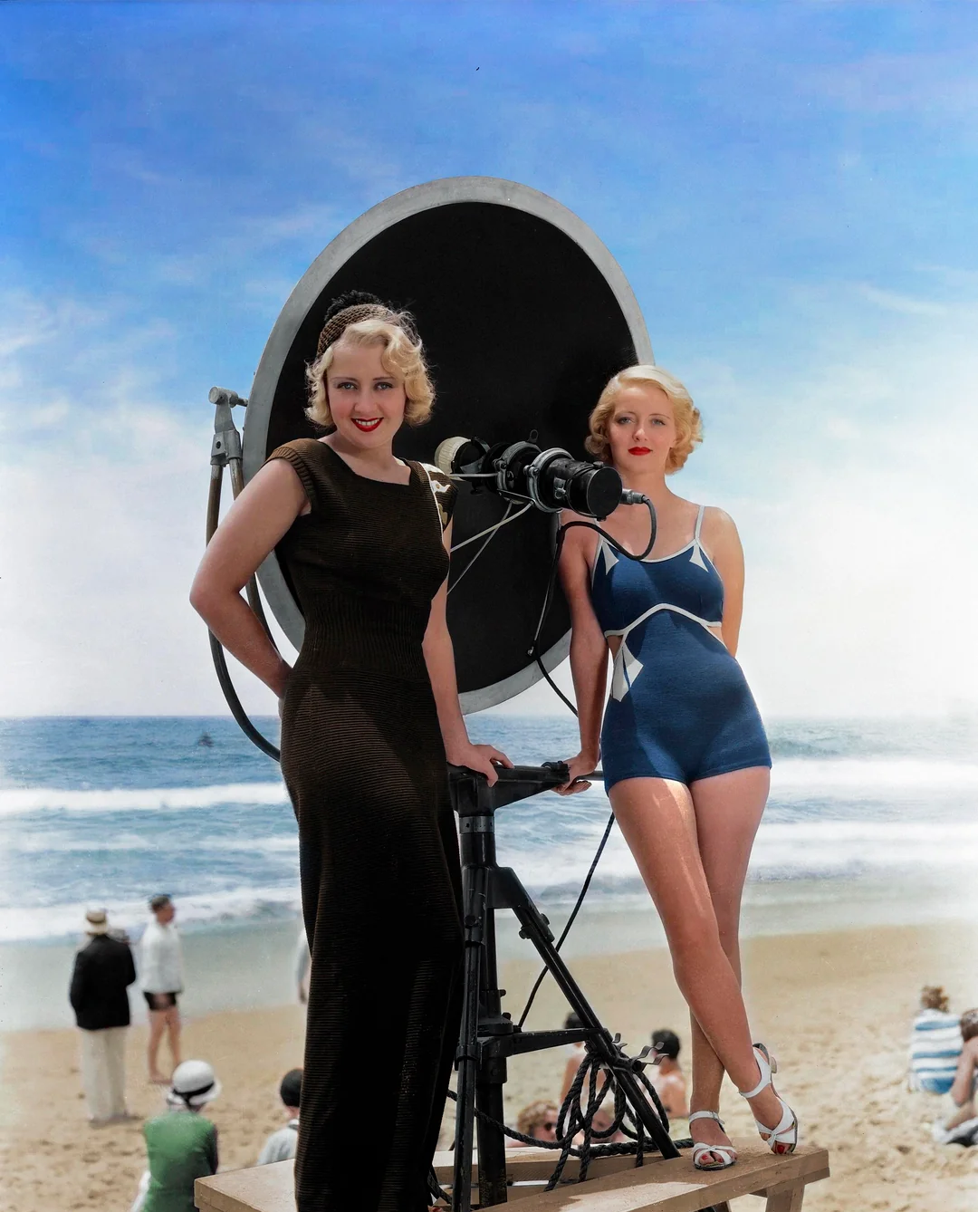 Two women with 1930s hairstyles stand by a large camera and reflector on a beach. One wears a dark dress; the other wears a blue swimsuit. The sea and people relaxing on the sand are visible in the background.