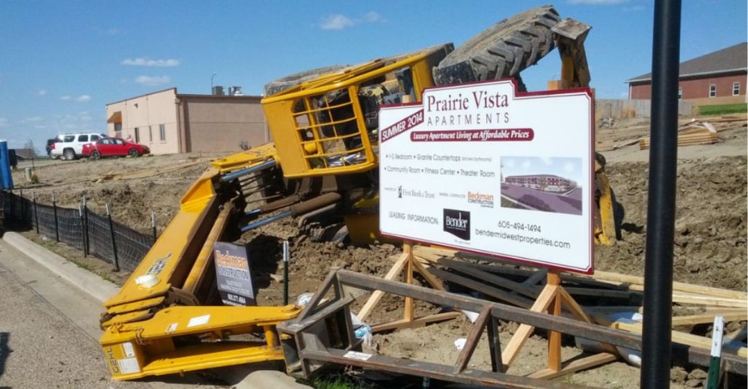 A yellow construction vehicle tipped over onto its side at a construction site near a Prairie Vista Apartments sign, with debris and disturbed dirt around the area.