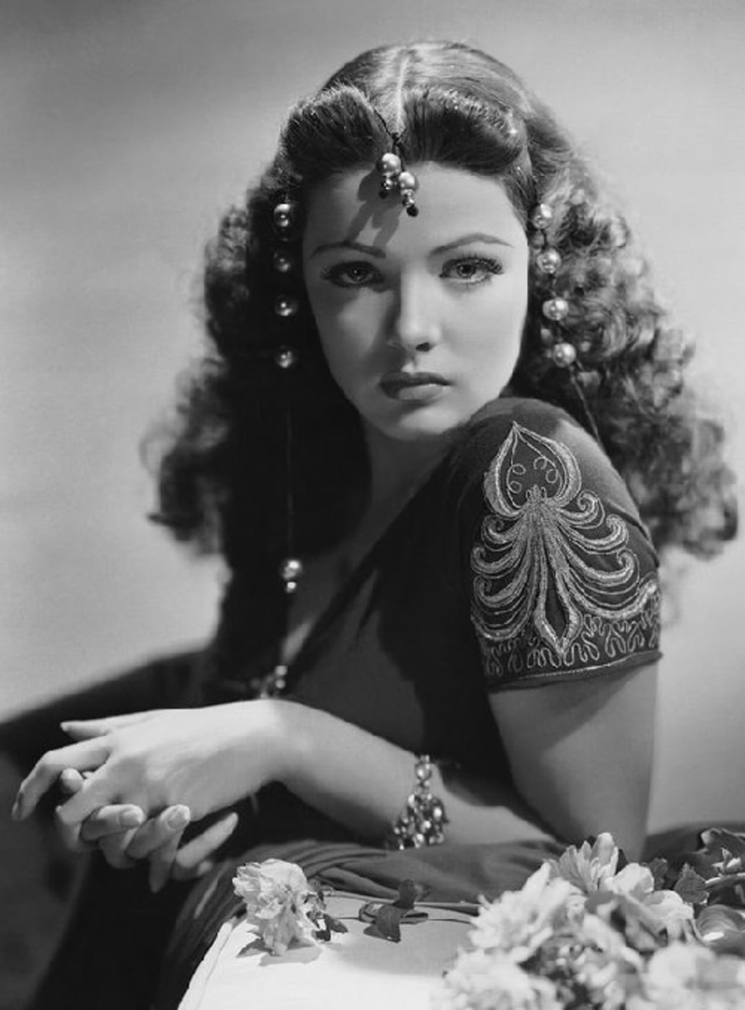A woman with long, wavy dark hair adorned with decorative beads poses with her hands together. She wears a short-sleeved dress with embroidered detail and a beaded bracelet, sitting beside flowers in a dramatic black-and-white portrait.