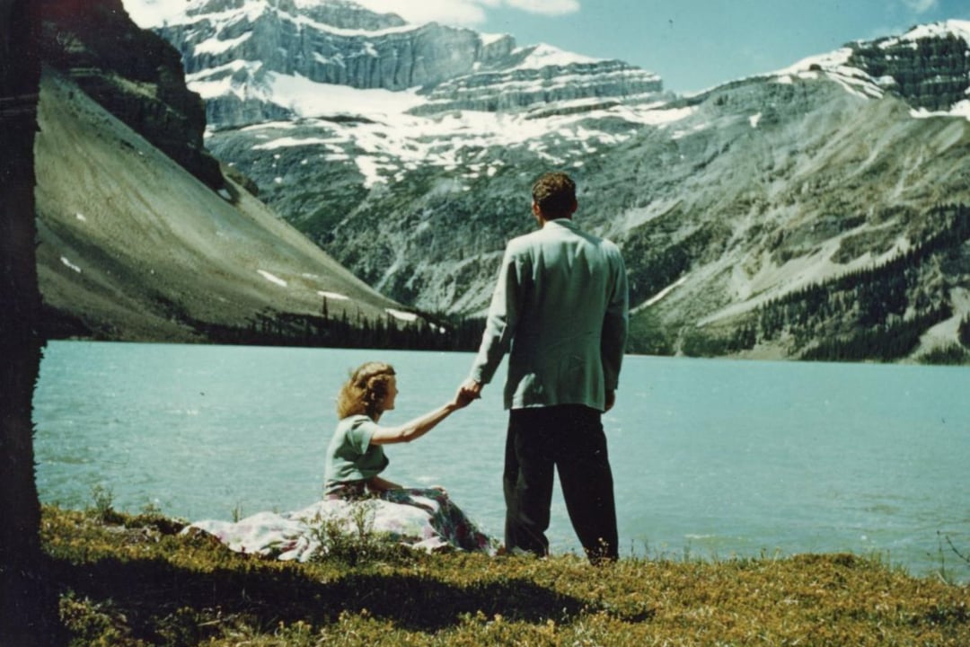 A woman sits on grass by a turquoise lake holding hands with a standing man. Snow-capped mountains and pine trees rise in the background under a partly cloudy sky.
