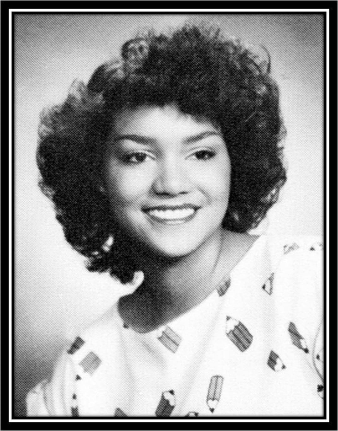 Black and white portrait of a young woman with curly hair, smiling and wearing a light-colored top with a repeating pencil pattern. The image has a thin black frame around it.