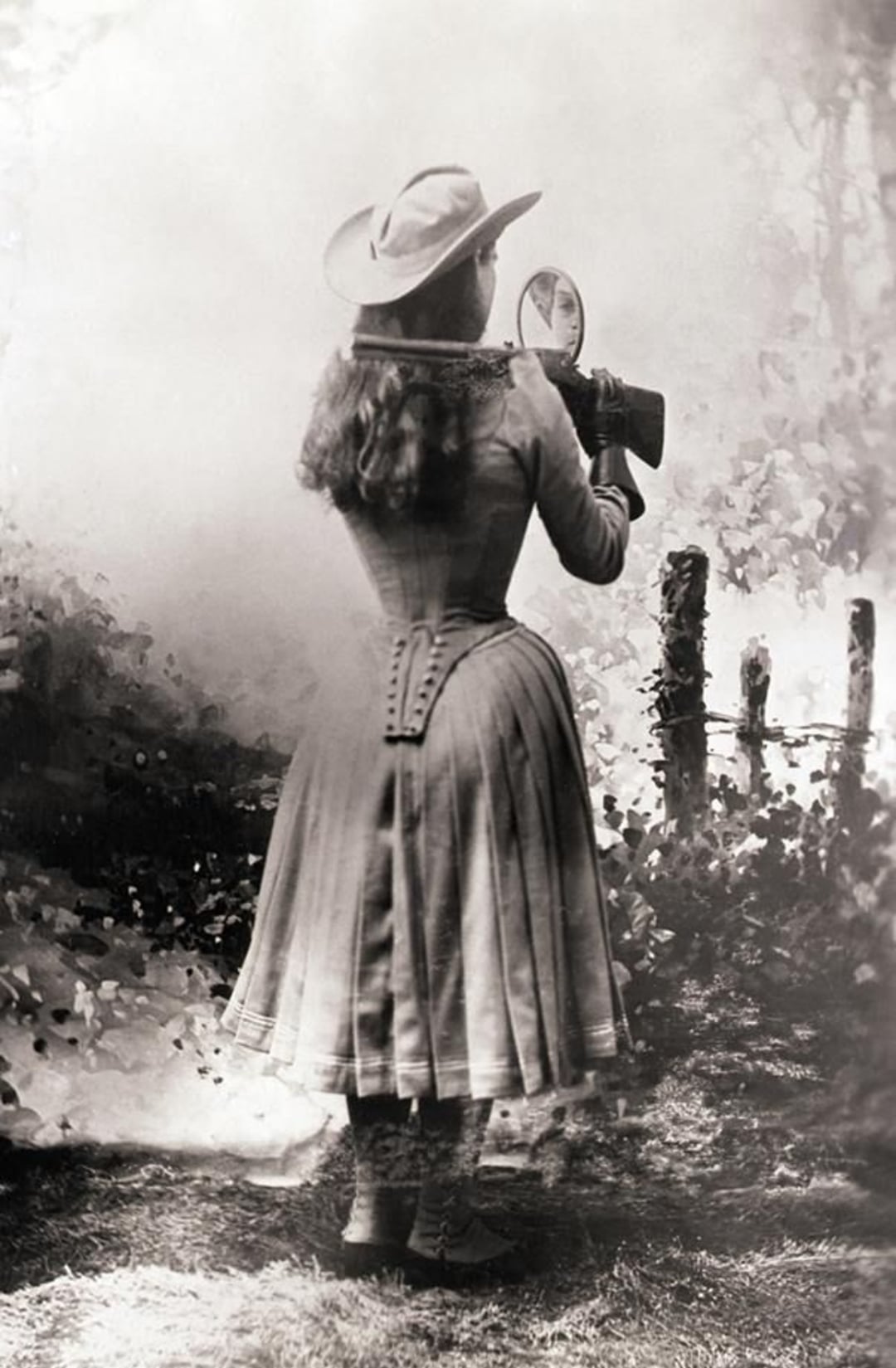 A woman in a long dress and wide-brimmed hat stands outdoors, aiming a rifle over her shoulder while looking into a small mirror for aiming, with a rustic fence and foliage in the background.