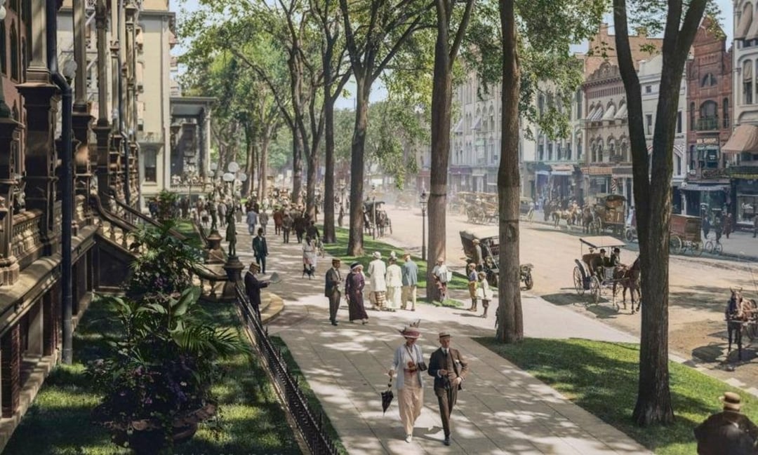 A busy city street scene from the early 1900s with people in period clothing walking on a tree-lined sidewalk, horse-drawn carriages on the road, and historic buildings lining both sides.