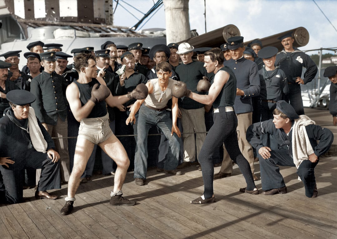 Two sailors wearing boxing gloves face off on the deck of a ship, surrounded by fellow sailors in uniforms who watch and cheer. Some sailors sit or lean on the railing, all appearing engaged in the match.