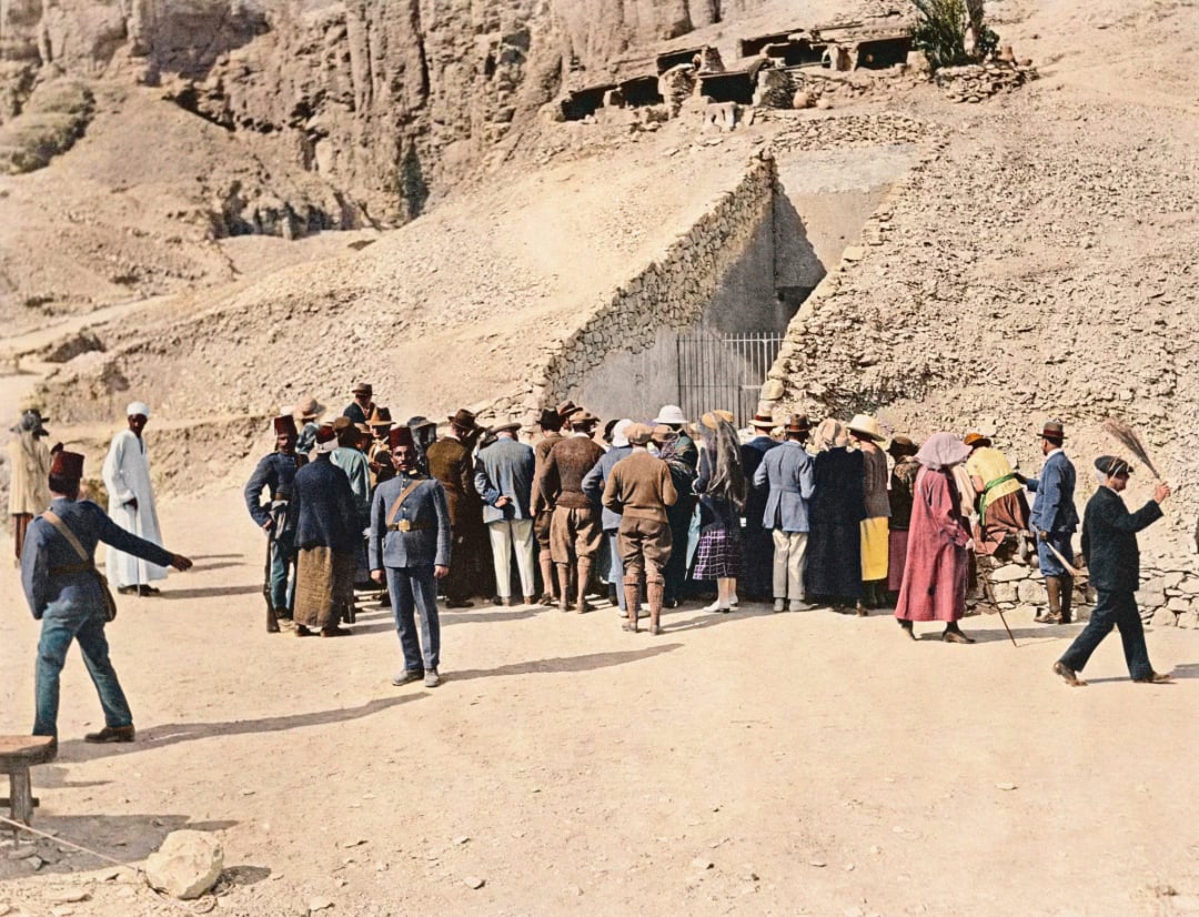 A group of people, including men in traditional clothing and pith helmets, gather at the entrance of an ancient stone tomb set in a rocky, desert landscape. Some guards stand nearby, and the area appears historical and archaeological.