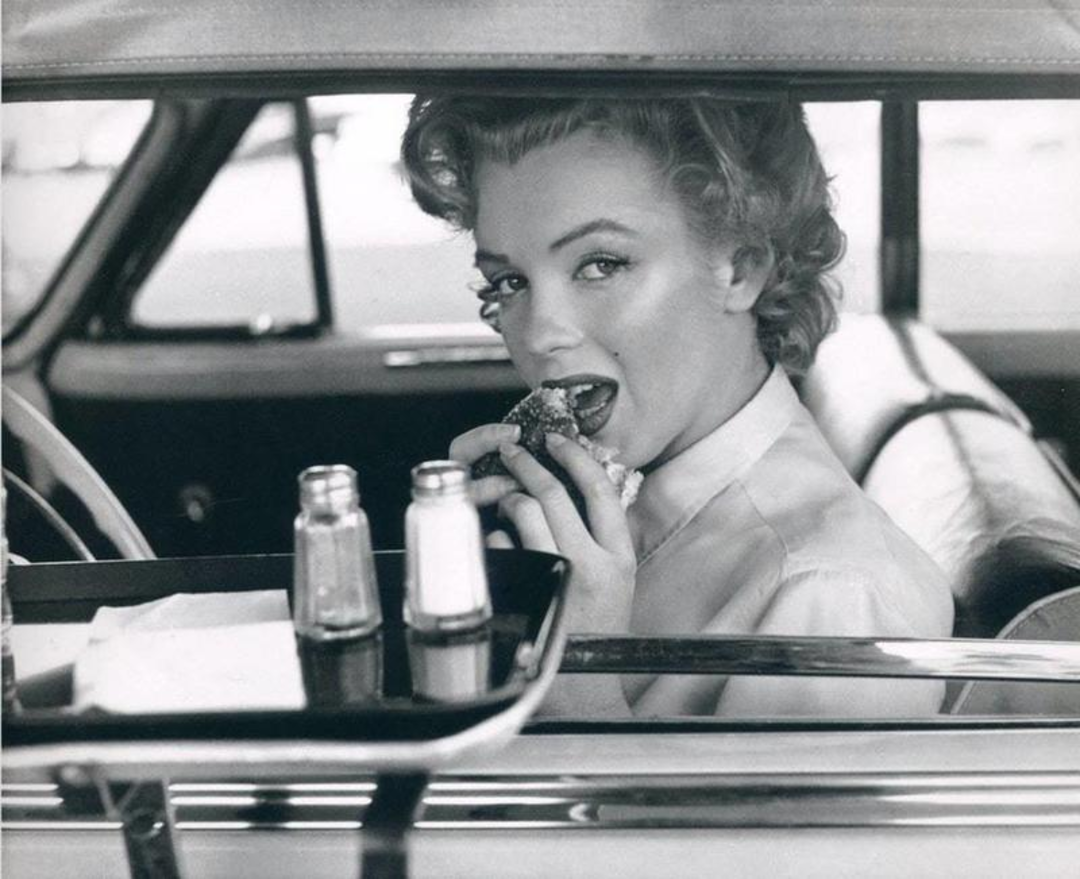 Black-and-white photo of a woman with styled hair sitting in a car, eating a hamburger. A food tray with salt and pepper shakers is attached to the car window. She looks toward the camera while taking a bite.