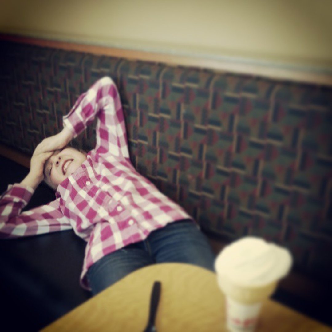 A person in a pink plaid shirt leans back on a cushioned bench, smiling with hands on their head. A table in the foreground holds a cup with a lid and a fork. The scene appears relaxed indoors.