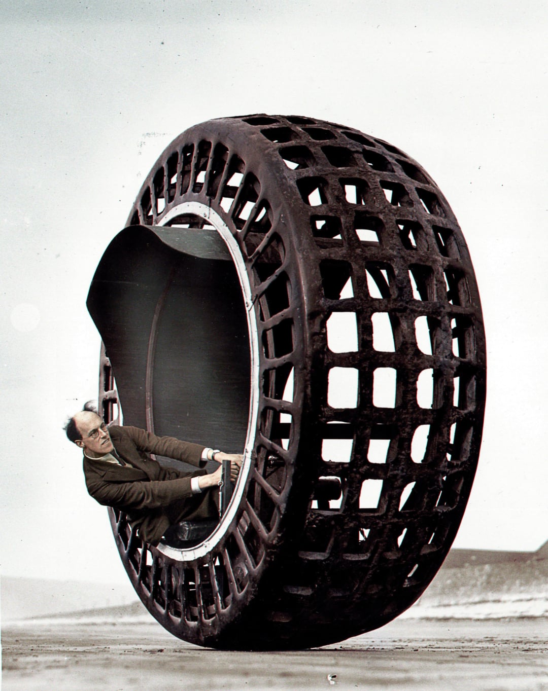 A man in a suit sits inside a large, hollow, lattice-structured wheel, appearing to steer or control it across a barren landscape. The scene has a surreal, vintage feel.