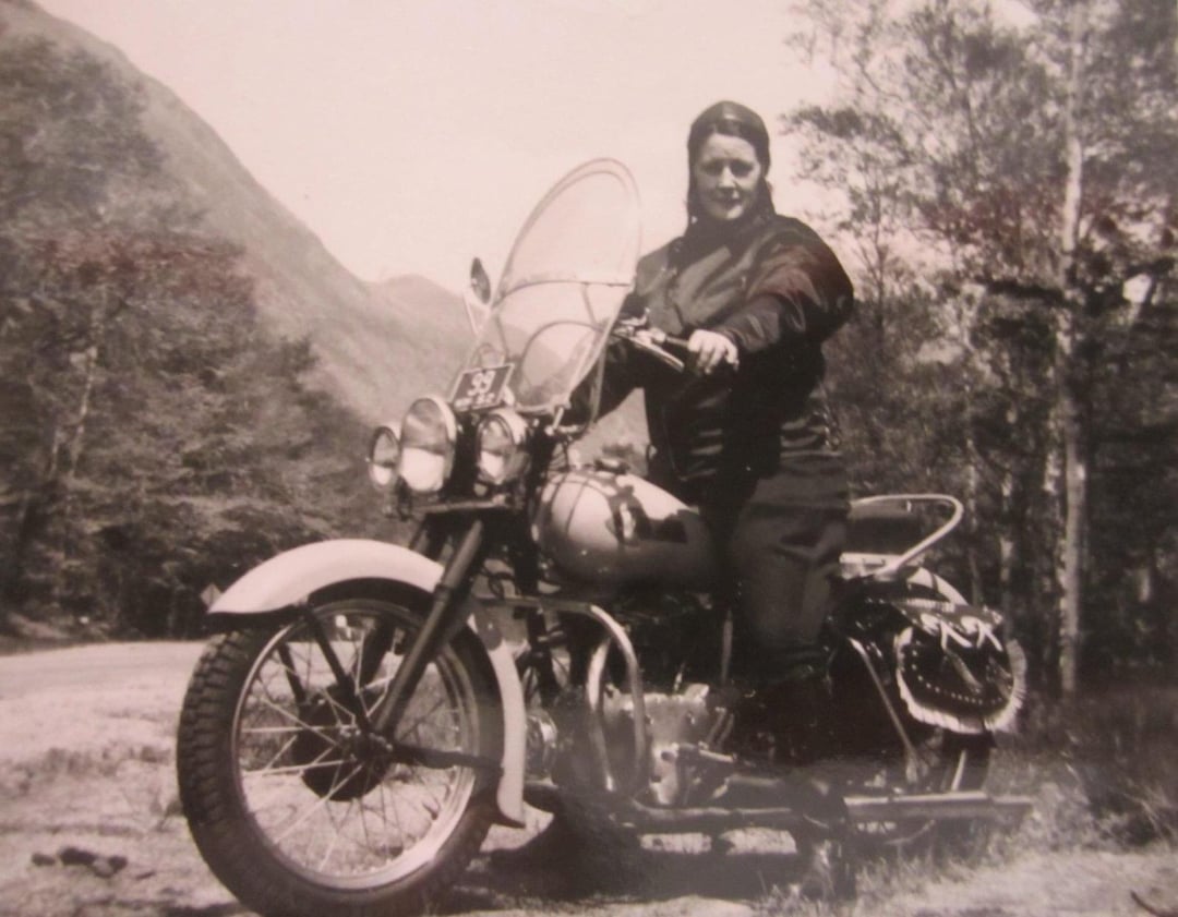 Black and white photo of a person in a jacket and headscarf sitting on a vintage motorcycle with a windshield, parked outdoors near trees and hills.