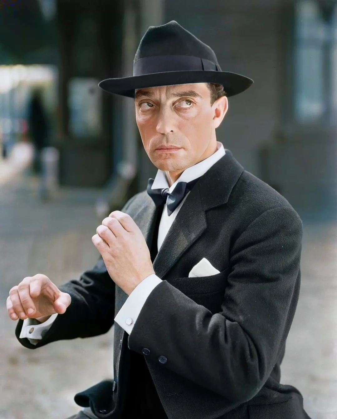 A man in a black suit, white shirt, and black bow tie stands with his hands raised. He wears a black hat and has a serious expression, standing outdoors near a building.
