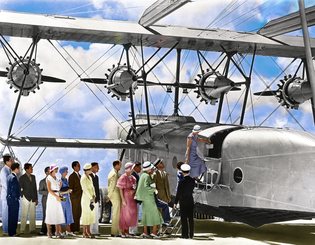 A group of well-dressed people in vintage clothing line up to board a large, silver, propeller-driven airplane with multiple engines, under a partly cloudy sky.