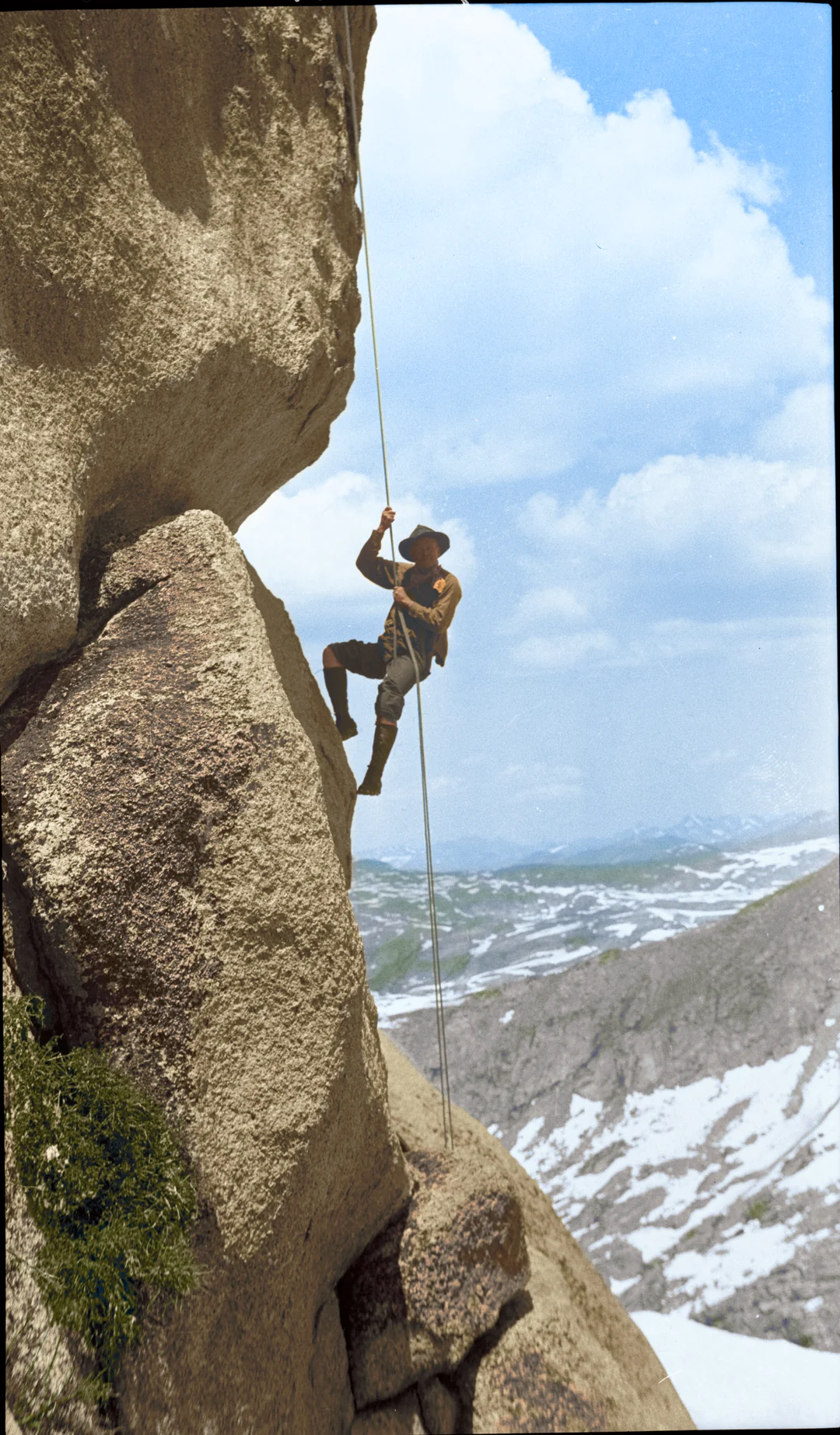 A person wearing outdoor gear and a wide-brimmed hat climbs a rope on a steep, rocky cliff with snow-capped mountains and a cloudy sky in the background.