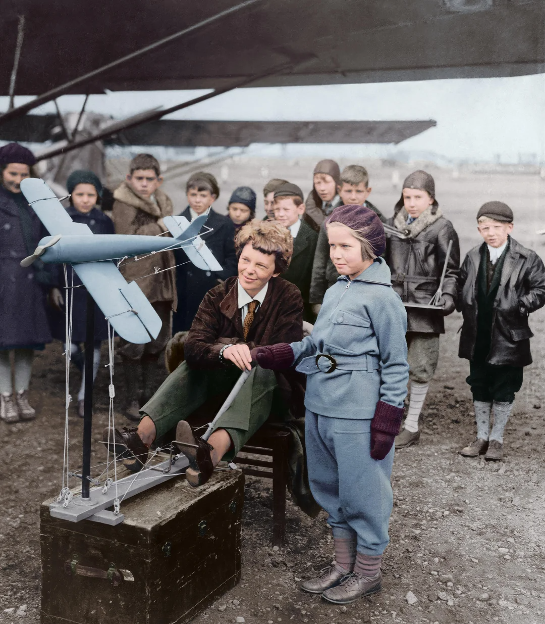 A group of children in vintage clothing gather outdoors around a model airplane. Two children stand closest, one seated operating the model, while others watch in the background, with a large real airplane behind them.