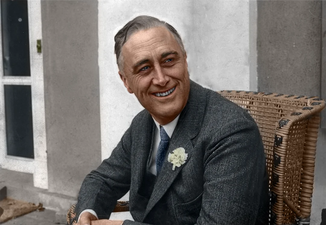 A man in a dark suit with a light-colored flower pinned to his lapel sits in a wicker chair, smiling. He has neatly combed hair and is positioned outdoors in front of a white wall.