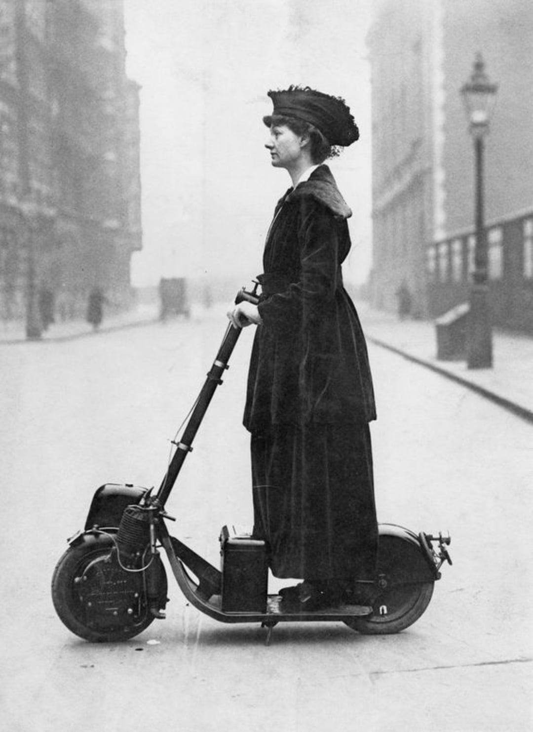 A woman in early 20th-century clothing stands on a vintage motorized scooter on a city street, with old buildings and a streetlamp visible in the background.