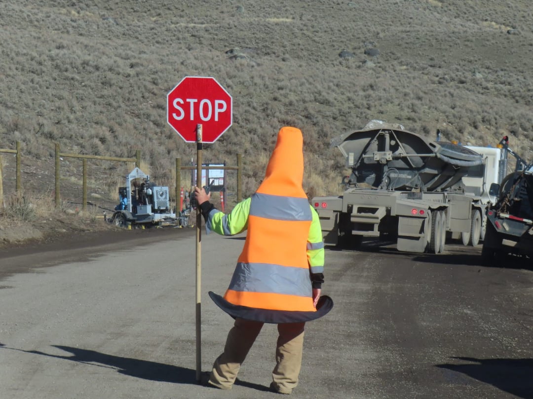 A person wearing a traffic cone costume and a reflective vest holds a stop sign on a rural dirt road, with construction vehicles and equipment in the background.