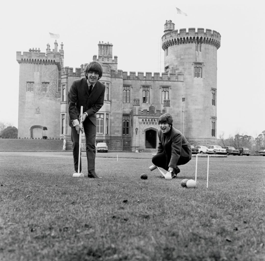 Two young men play croquet on a lawn in front of a large stone castle with towers. Both are smiling and dressed in suits. Several cars are parked near the castle in the background.