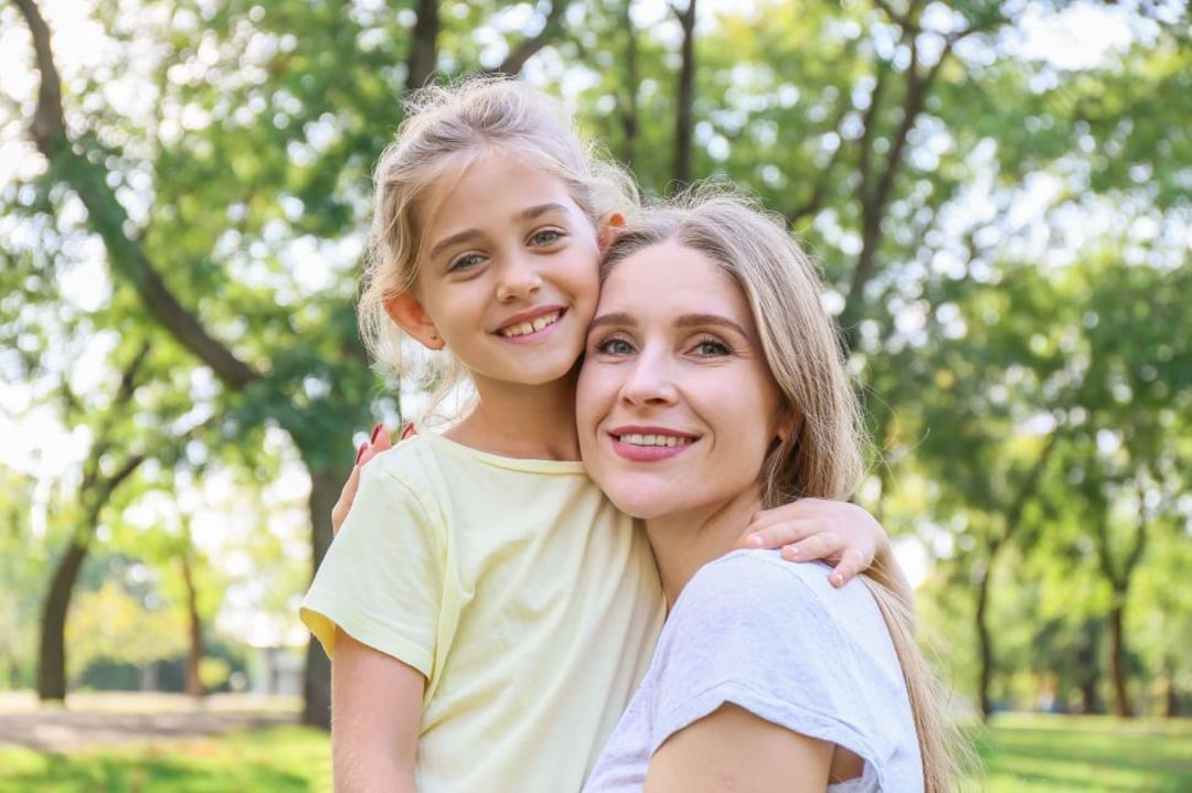 A smiling woman and young girl embrace outdoors in a sunlit park, with green trees and grass in the background. The girl wears a yellow shirt and the woman wears a white shirt. Both look happy.