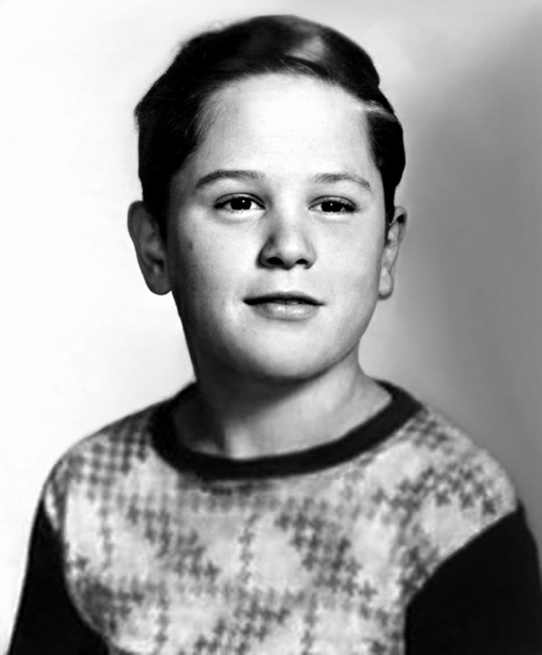 A black-and-white portrait of a young boy with short, neatly combed hair, wearing a patterned sweater. He is looking slightly to the side with a neutral expression against a plain background.