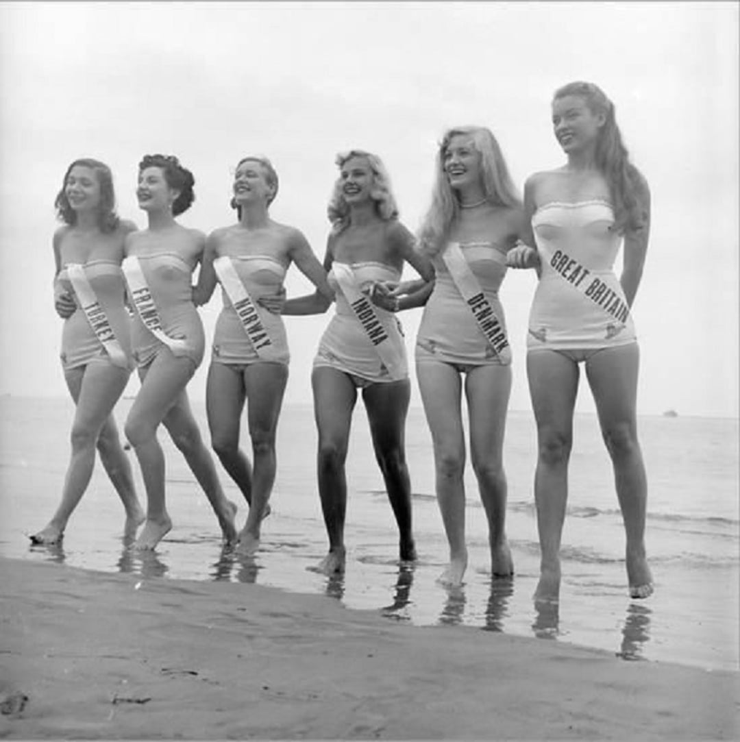 Six women in swimsuits, each wearing a sash with a country name, stand side by side on a beach, smiling and holding arms. The sashes read USA, Chile, France, Finland, Denmark, and Great Britain.