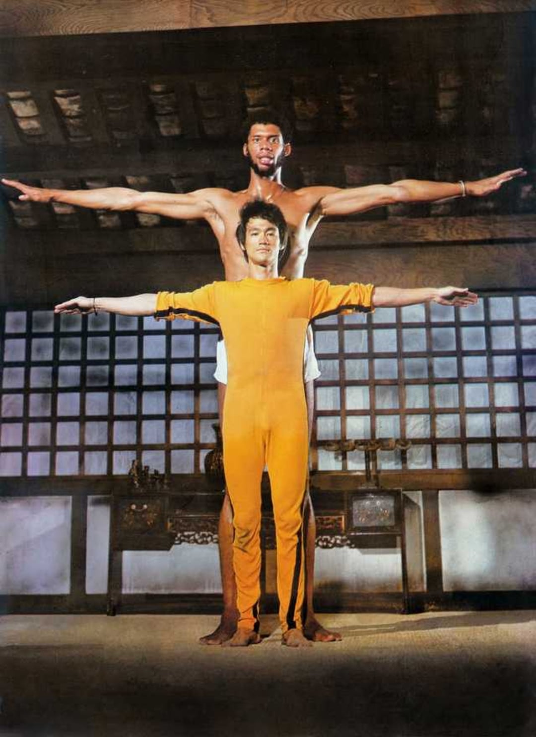 Two men stand barefoot indoors with their arms outstretched horizontally, one in a yellow jumpsuit in front and a much taller man behind, both facing forward against a backdrop of wooden beams and shoji-style windows.