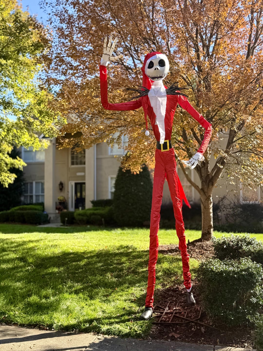 A tall Jack Skellington figure in a red Santa suit stands in a front yard, waving. Autumn trees with orange leaves and a large house are in the background, and sunlight casts shadows on the grass.
