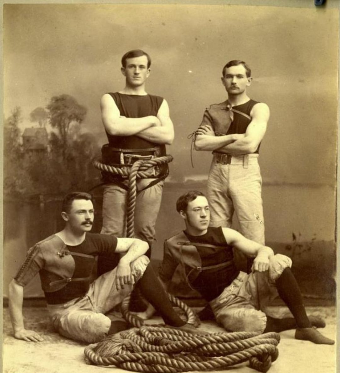 Four men in vintage athletic uniforms posing with large ropes. Two are standing with arms crossed, and two are seated on the floor. The background features a painted outdoor scene. All have mustaches and serious expressions.