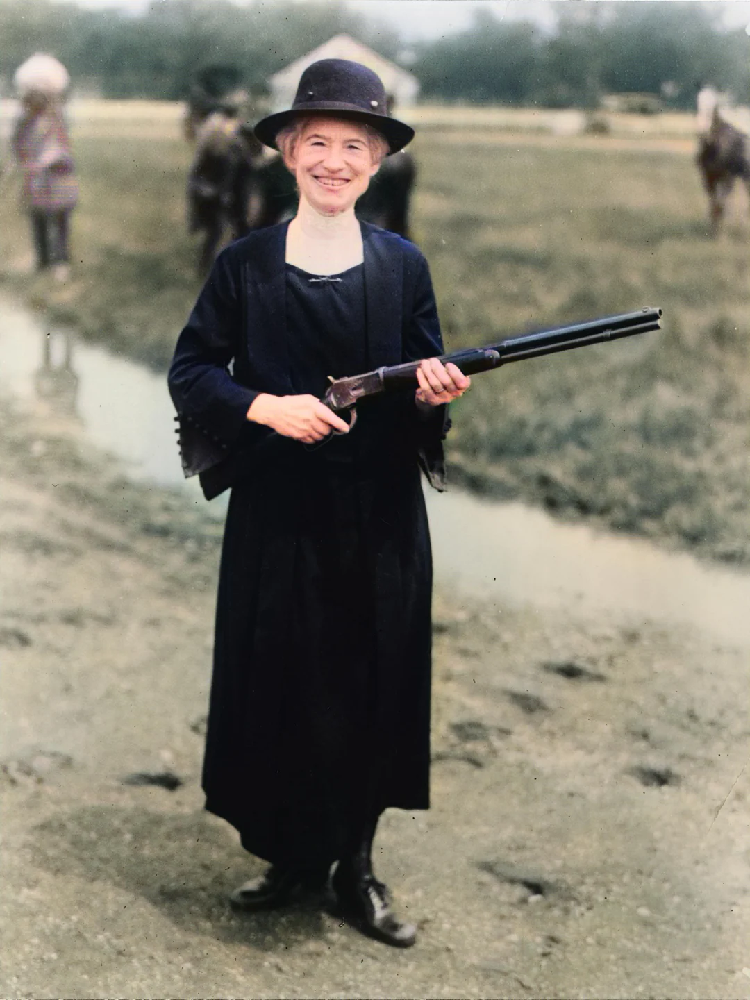 A smiling woman in a dark dress, boots, and hat stands on a muddy path holding a shotgun. The background shows blurred people, horses, and green fields.