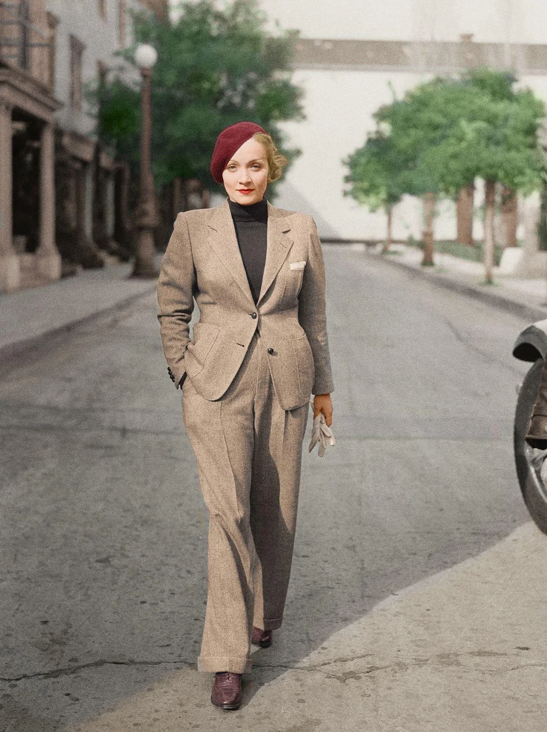 A person wearing a tan suit, black turtleneck, red beret, and brown shoes walks confidently down a quiet, tree-lined street holding white gloves.