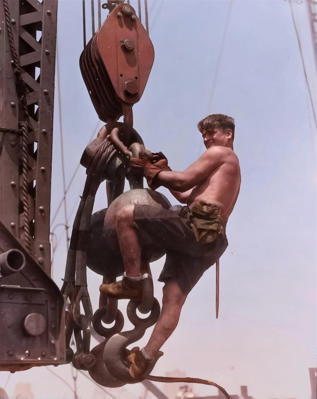 A shirtless male construction worker wearing shorts, boots, and gloves hangs onto a large metal hook and pulley of a crane, high above the ground, with ropes and machinery in the background.