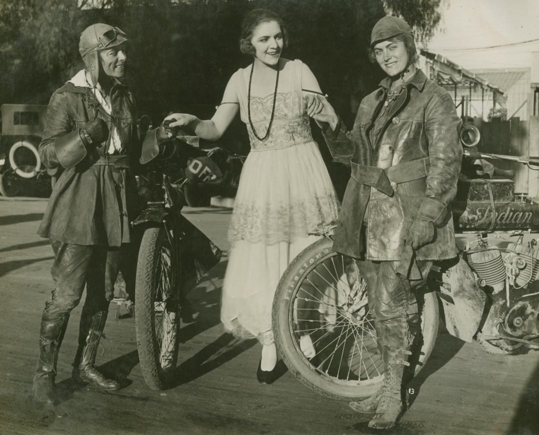 Two women in leather motorcycle gear stand with their bikes, smiling, while a woman in a dress and pearls stands between them, holding their arms. Vintage cars are visible in the background.