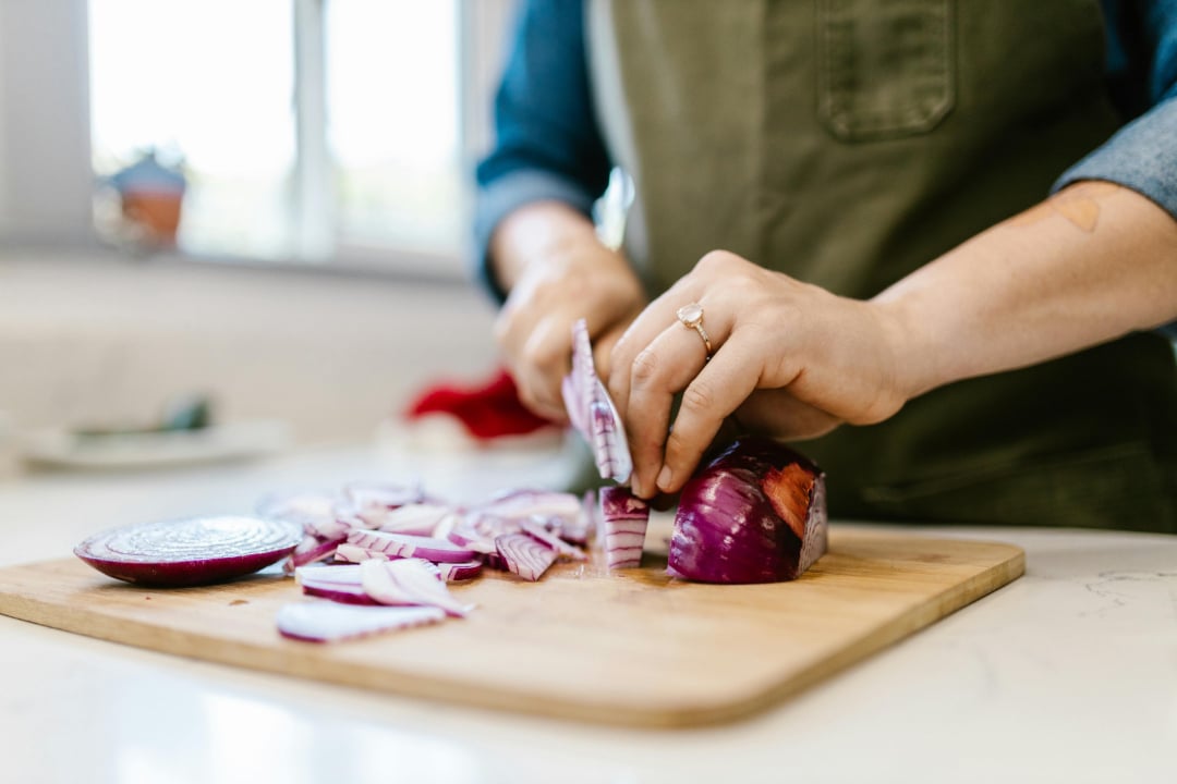 A person wearing an apron slices a red onion on a wooden cutting board in a bright kitchen. Sliced onion pieces are scattered on the board.