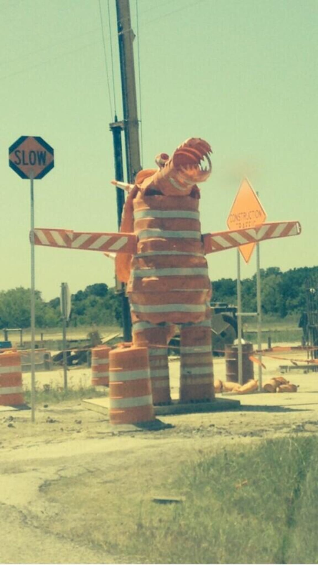 A large dinosaur sculpture made from orange construction barrels and cones stands at a roadside construction site, with "Slow" and "Construction Ahead" signs nearby.